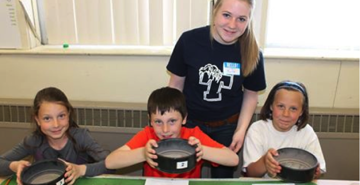 A young female science student standing behind a groupp of three young students while they work on a science project